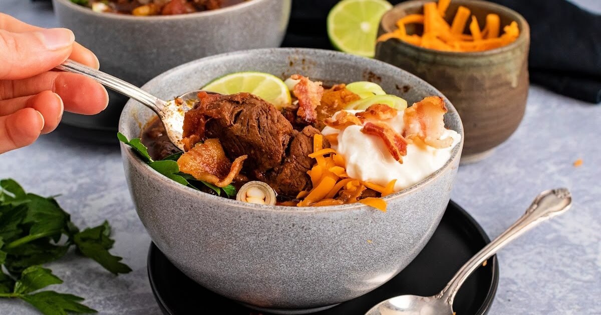 Bowl full of meat chili with chunks of stew meat, topped with shredded cheddar cheese, sour cream, bacon pieces, green onions, fresh cilantro and a lime wedge, sitting on plates with a spoon next to it and a hand scooping a spoonful of chili out of the bowl, with a small bowl of shredded cheese and a lime wedge in the background.