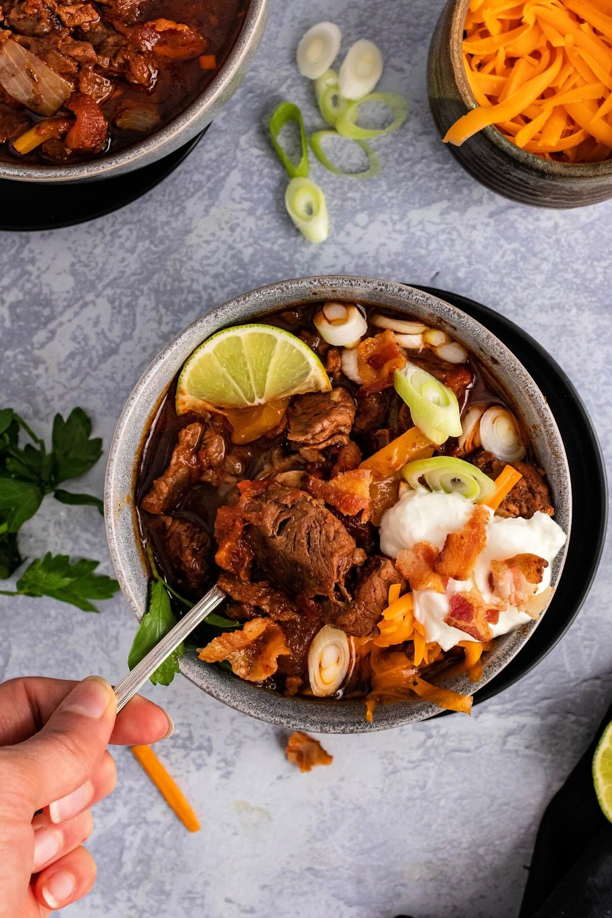 Bowl full of meat chili with chunks of stew meat, topped with shredded cheddar cheese, sour cream, bacon pieces, green onions, fresh cilantro and a lime wedge, sitting on a plate with a hand scooping a spoonful of chili out of the bowl, with a small bowl of shredded cheese, fresh cilantro spring, chopped green onions and another bowl of chili in the background.