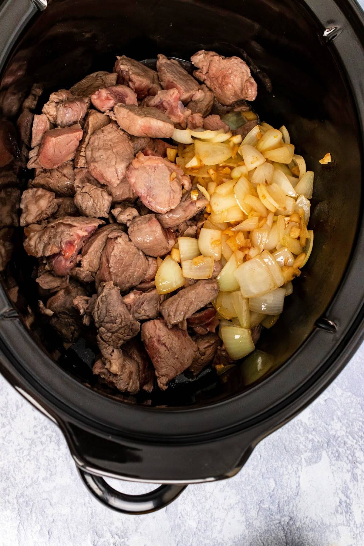 Slow cooker filled with browned stew meat and caramelized onions.