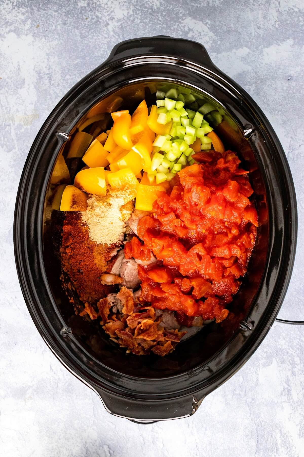 Slow cooker filled with canned tomatoes, chopped yellow bell peppers, chopped celery, chopped cooked bacon and lots of spices and herbs on top of browned stew meat.