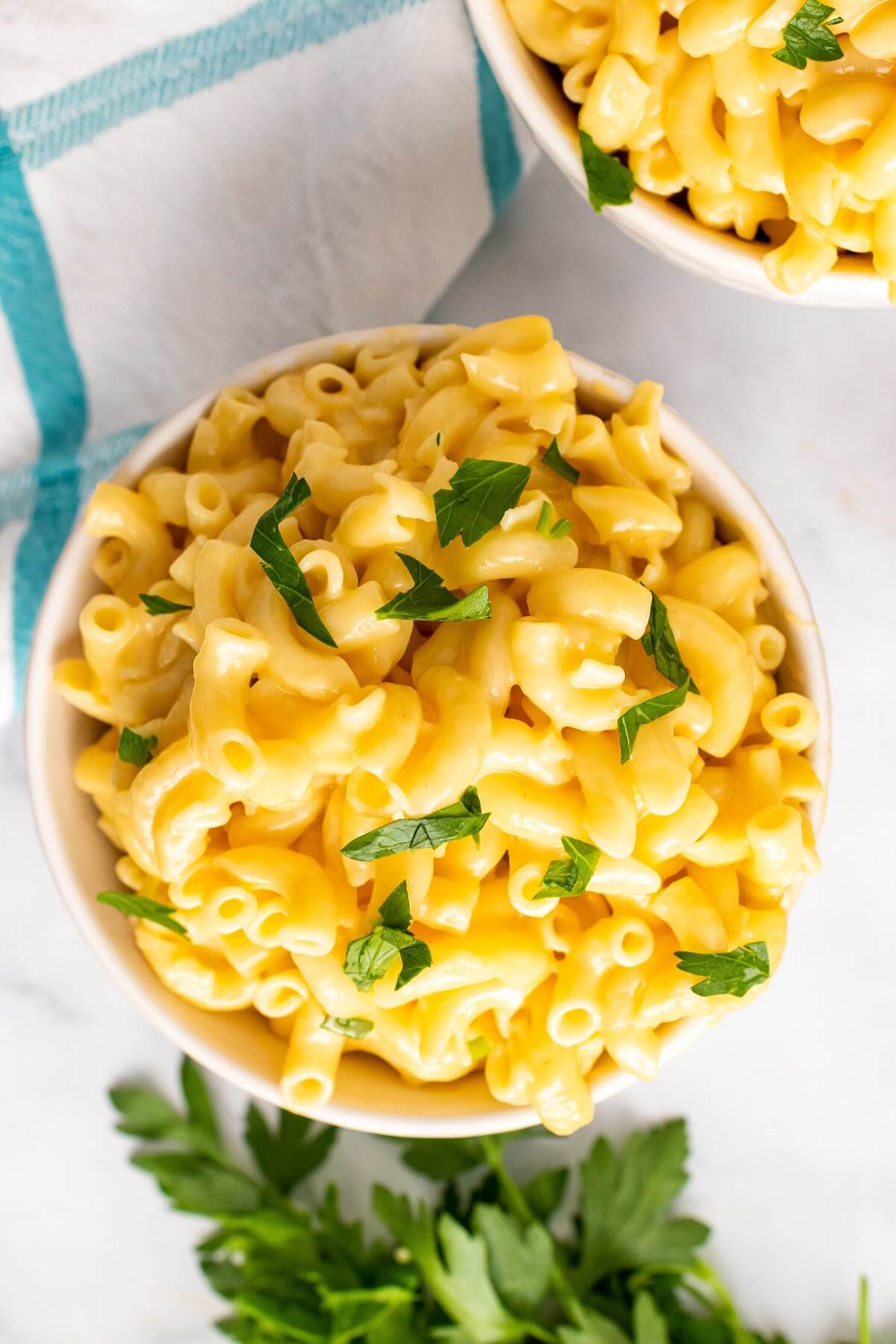Two bowls full of homemade Mac and cheese, garnished with fresh parsley sitting next to fresh Italian parsley sprigs and a cloth kitchen towel.