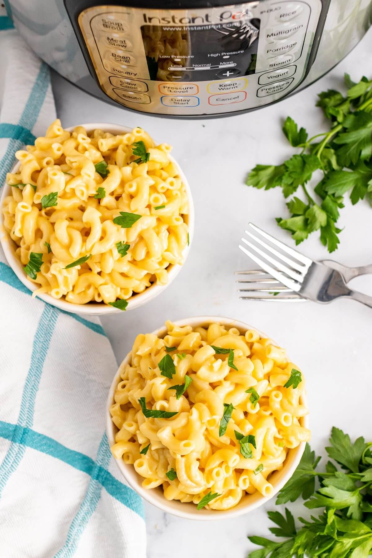 Two bowls full of homemade Mac and cheese, garnished with fresh parsley sitting next to two forks, fresh Italian parsley sprigs, a cloth kitchen towel and an Instant Pot.