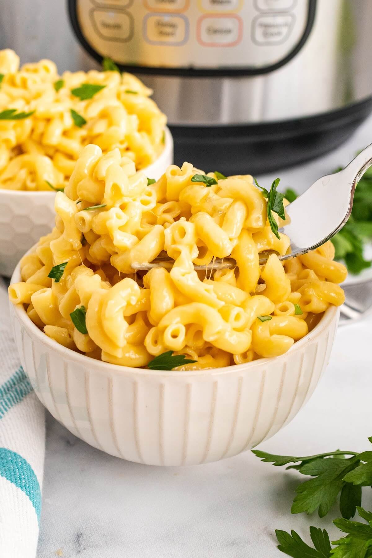 Two bowls full of homemade Mac and cheese, one with a fork dipping into the Mac and cheese, garnished with fresh parsley sitting next to fresh Italian parsley sprigs, a cloth kitchen towel and an Instant Pot.