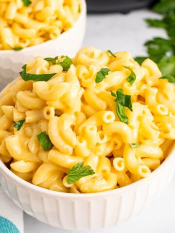 Two bowls full of homemade Mac and cheese, garnished with fresh parsley sitting next to two forks, fresh Italian parsley sprigs, a cloth kitchen towel and the base of an Instant Pot in the background.