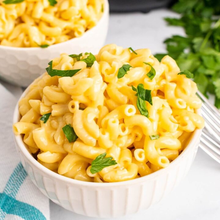 Two bowls full of homemade Mac and cheese, garnished with fresh parsley sitting next to two forks, fresh Italian parsley sprigs, a cloth kitchen towel and the base of an Instant Pot in the background.