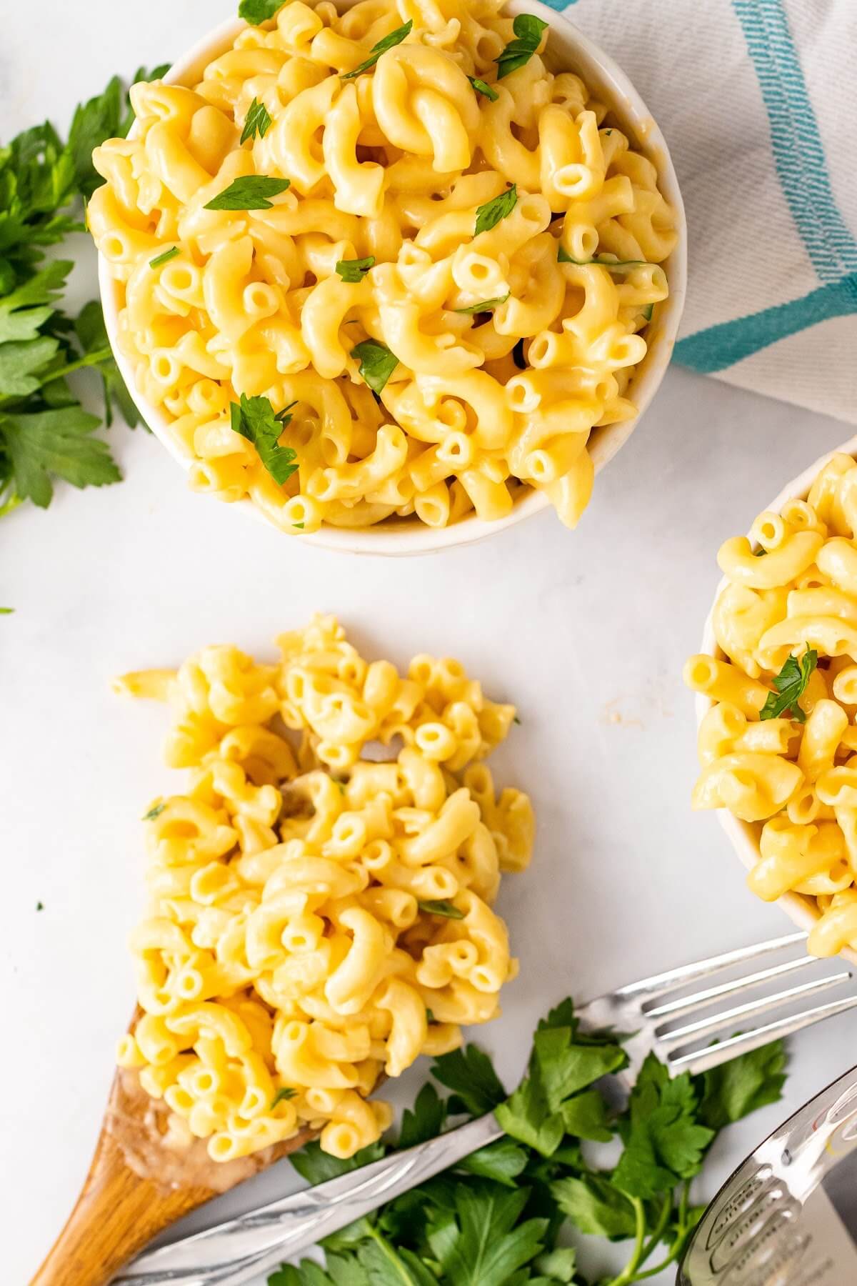 Two bowls full of homemade Mac and cheese, garnished with fresh parsley sitting next to a fork, a wooden spoon full of Mac and cheese, fresh Italian parsley sprigs, a cloth kitchen towel and an Instant Pot.
