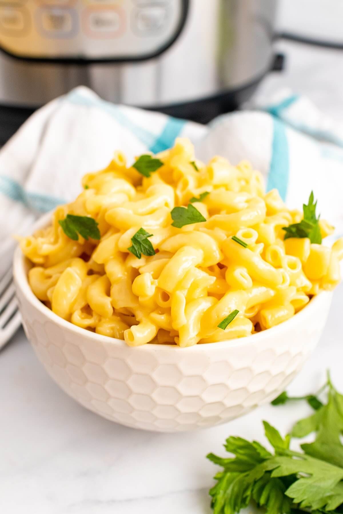 One bowl full of homemade Mac and cheese, garnished with fresh parsley sitting next to a fork, fresh Italian parsley sprigs, a cloth kitchen towel and an Instant Pot.