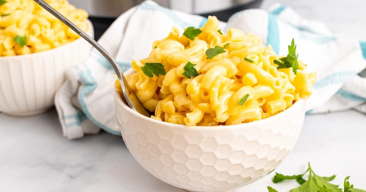 Two bowls full of homemade Mac and cheese, one with a fork in it, garnished with fresh parsley sitting next to fresh Italian parsley sprigs, a cloth kitchen towel and an Instant Pot.