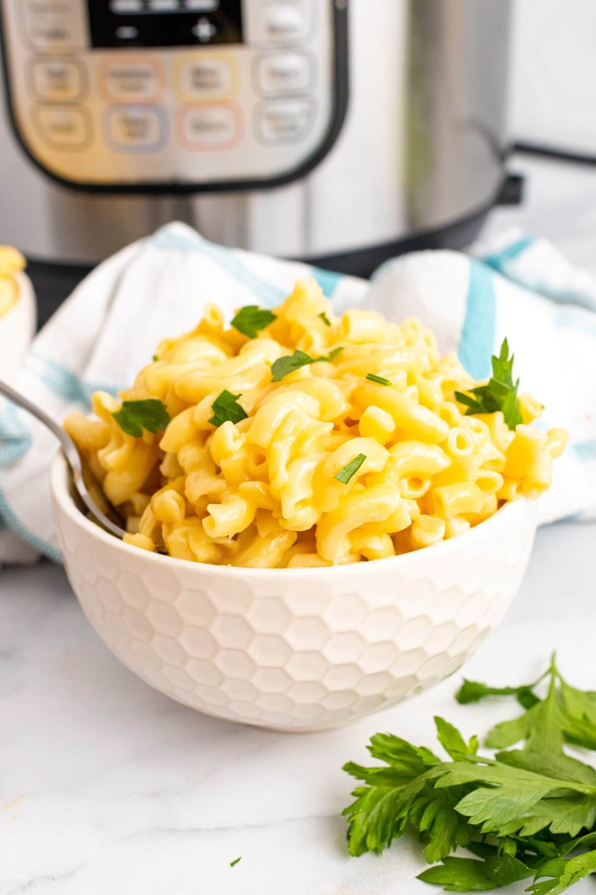 One bowl full of homemade Mac and cheese with a fork in it, garnished with fresh parsley sitting next to fresh Italian parsley sprigs, a cloth kitchen towel and an Instant Pot.