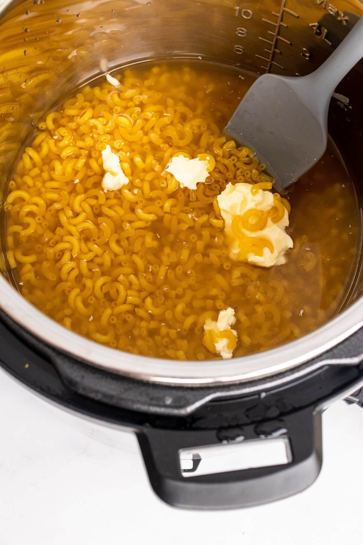 An Instant Pot filled with dry macaroni pasta, broth and butter being stirred with a spatula.