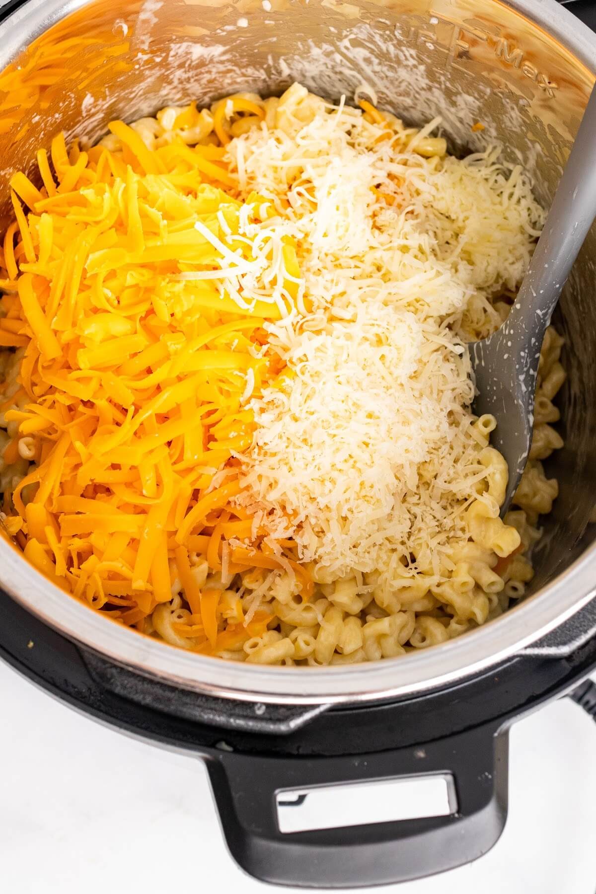 An Instant Pot filled with cooked macaroni pasta and topped with several kinds of shredded cheeses being stirred with a spatula.