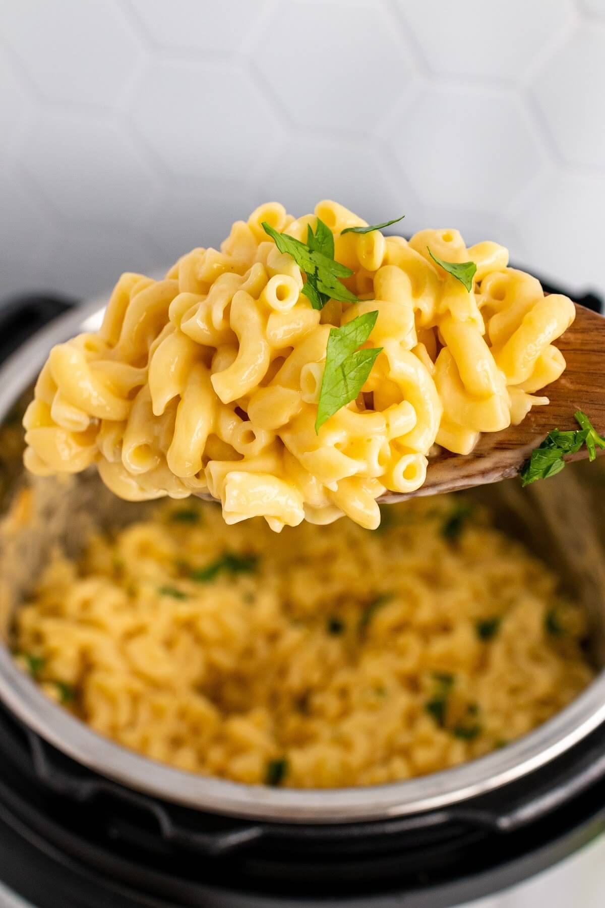 A wooden spoon full of homemade Mac and cheese above an Instant Pot filled with cooked homemade Mac and cheese garnished with fresh parsley.