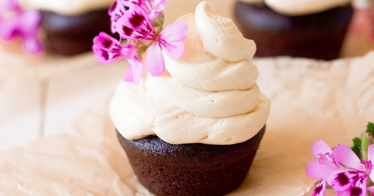 Chocolate cupcake topped with a 2-inch swirl of vanilla buttercream frosting and pink geranium flowers for garnish sitting on top of a square piece of unbleached parchment paper and two vanilla frosted cupcakes in the background.
