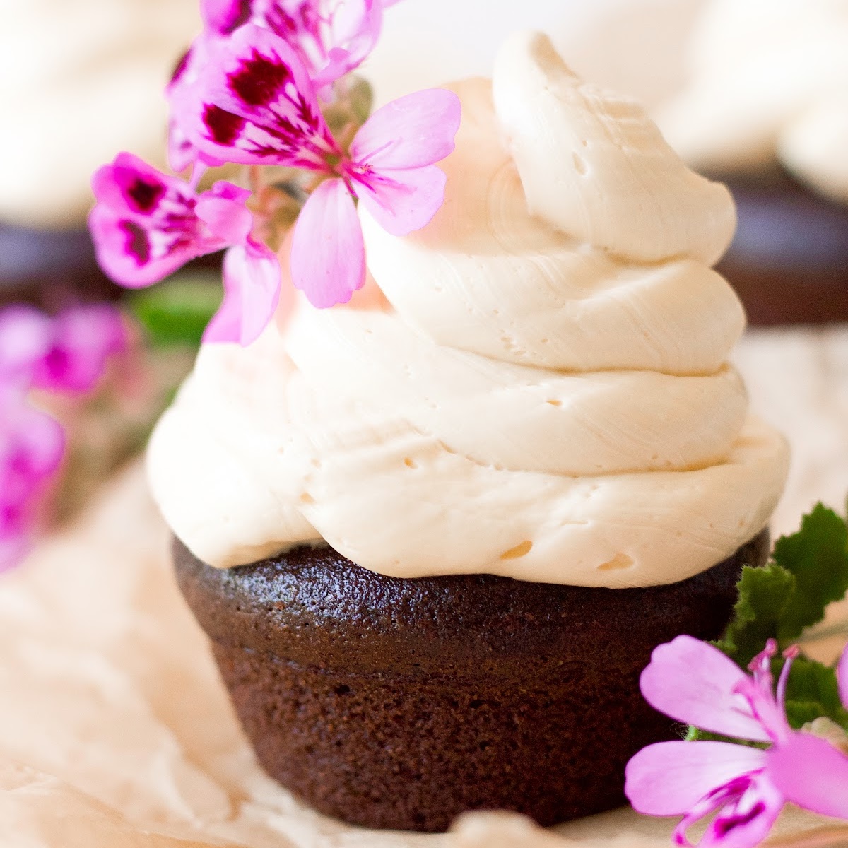 Chocolate cupcake topped with a 2-inch swirl of vanilla buttercream frosting and pink geranium flowers for garnish.