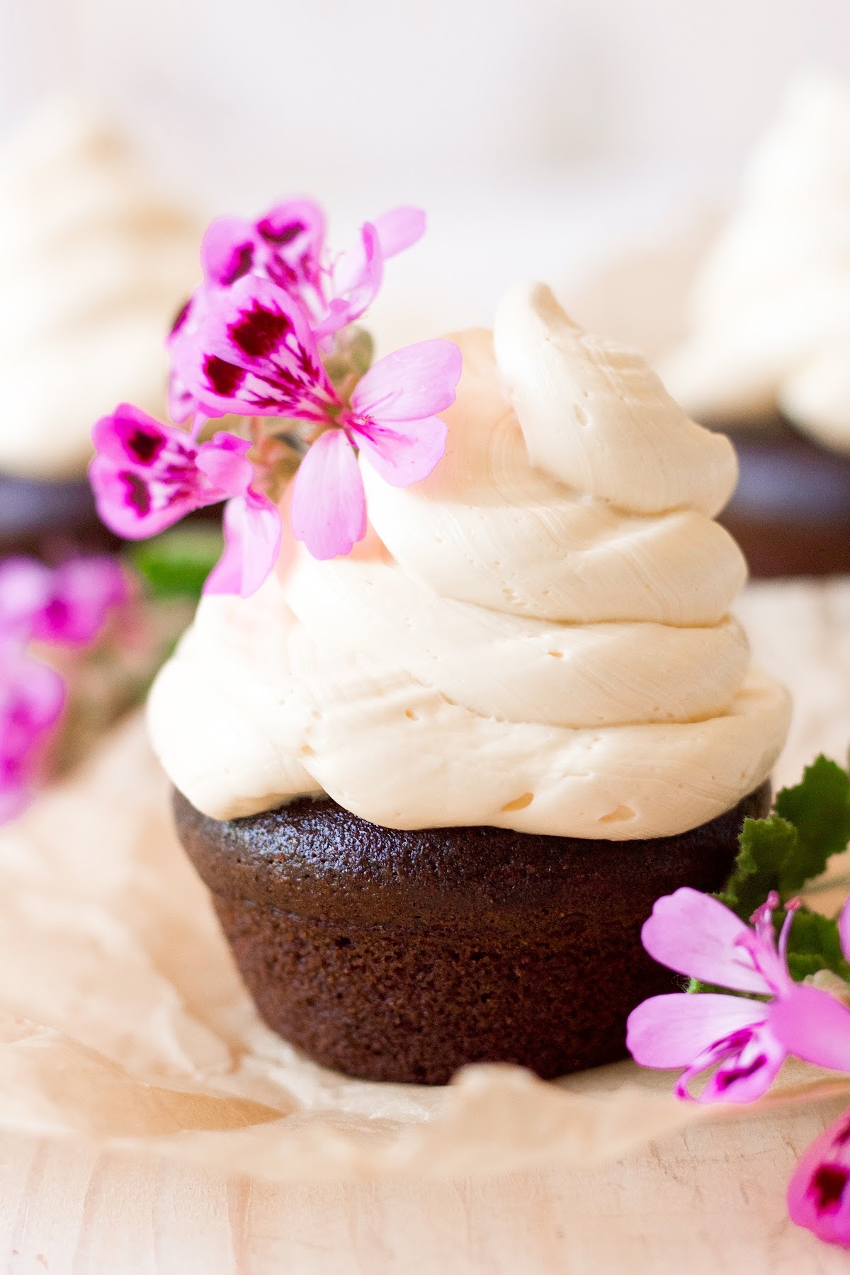 Chocolate cupcake topped with a 2-inch swirl of vanilla buttercream frosting and pink geranium flowers for garnish sitting on top of a square piece of unbleached parchment paper.