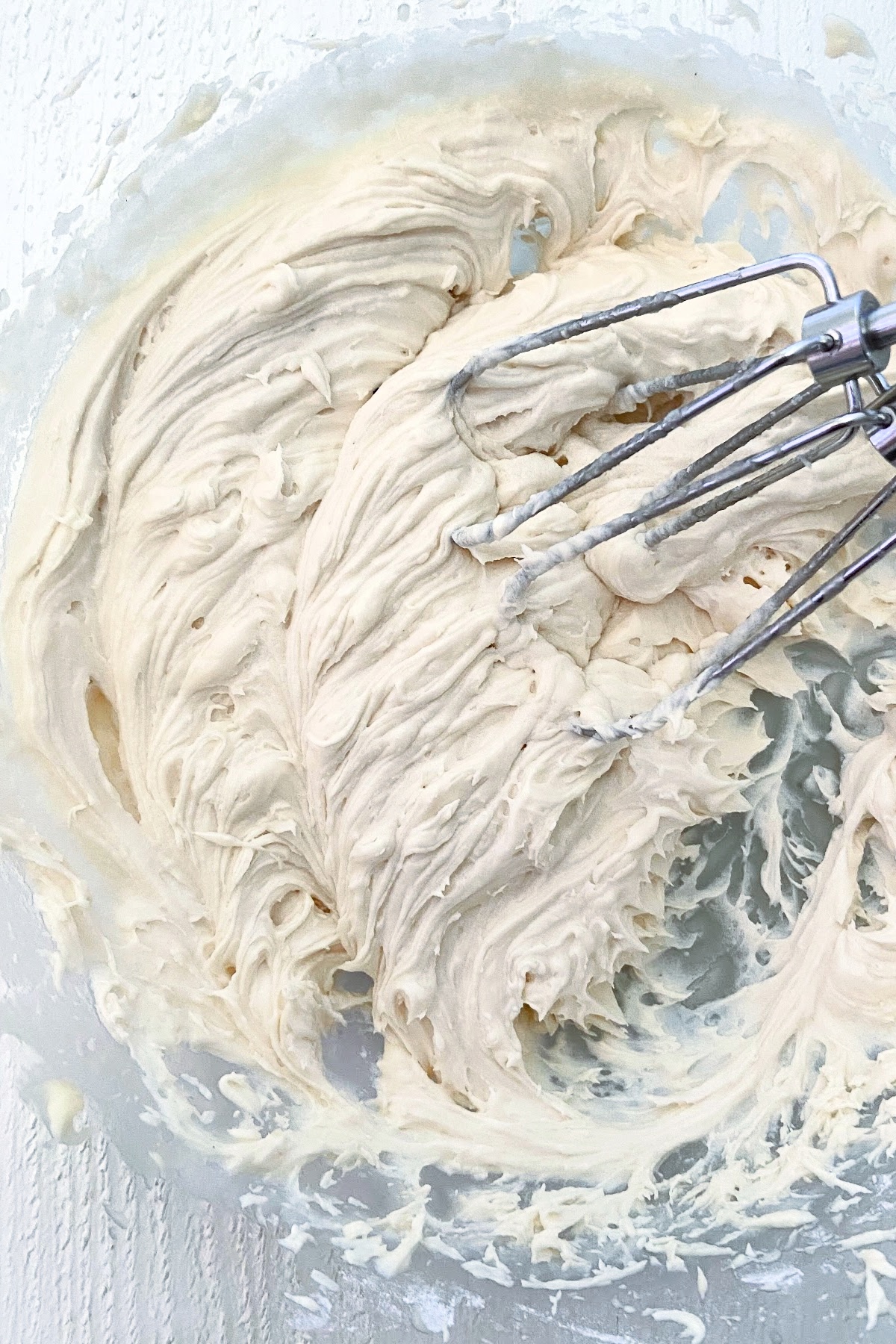 Close up photo of a glass mixing bowl filled with whipped vanilla buttercream frosting with 2 handheld mixer beaters mixing the frosting.