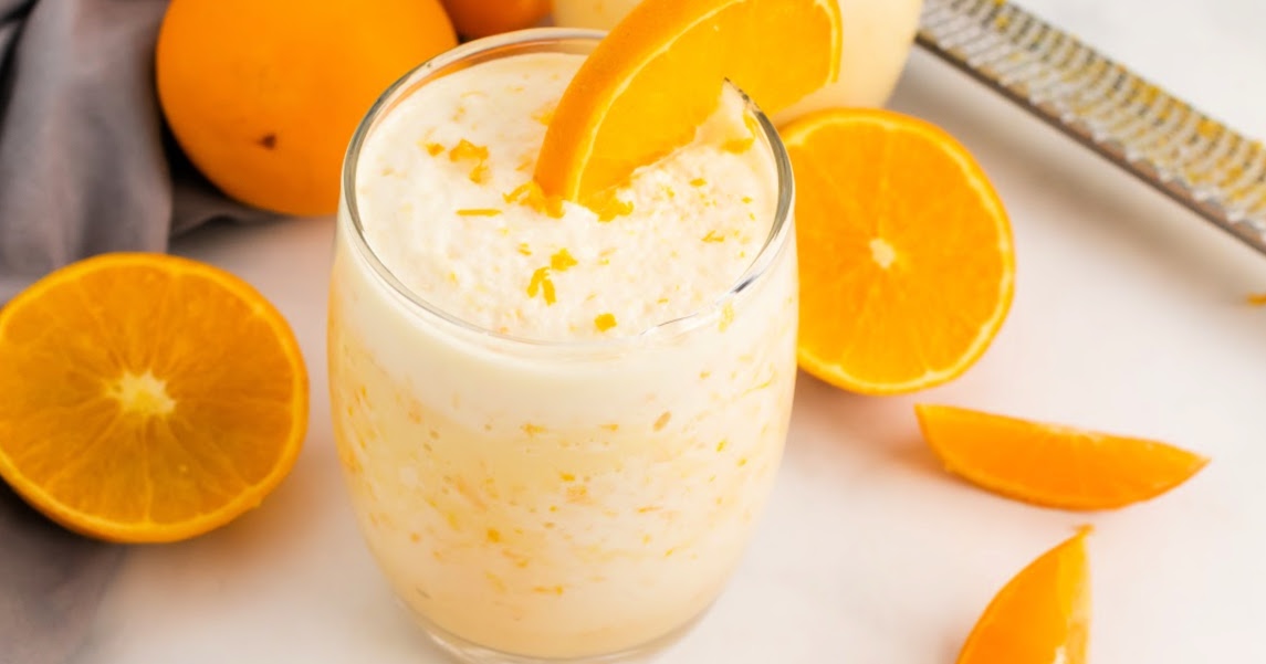 Glass full or a creamy orange smoothie with a microplane sitting in the background with oranges and orange slices.