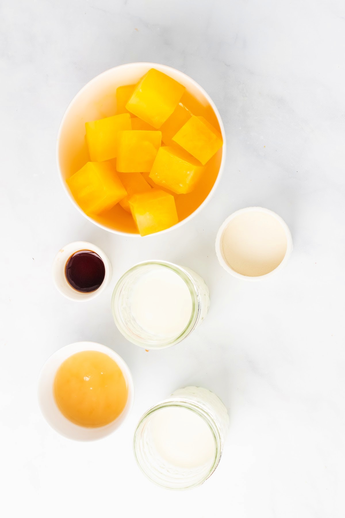 Bowl full of frozen orange juice ice cubes, small bowl with honey, small bowl with vanilla extract, small glass jar with milk, small glass jar with cream and small bowl with hydrolyzed collagen.