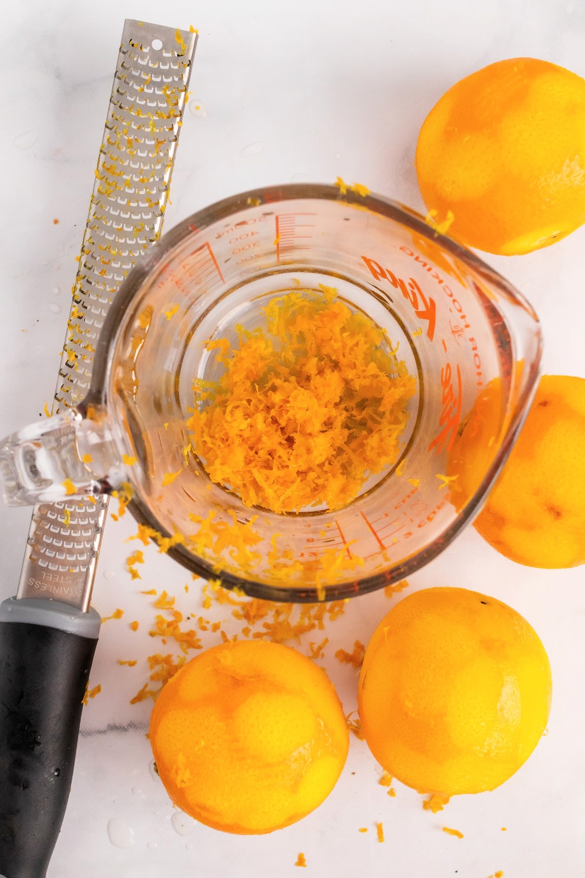Glass measuring cup filled with orange zest with a microplane sitting next to it and 4 zested whole oranges.