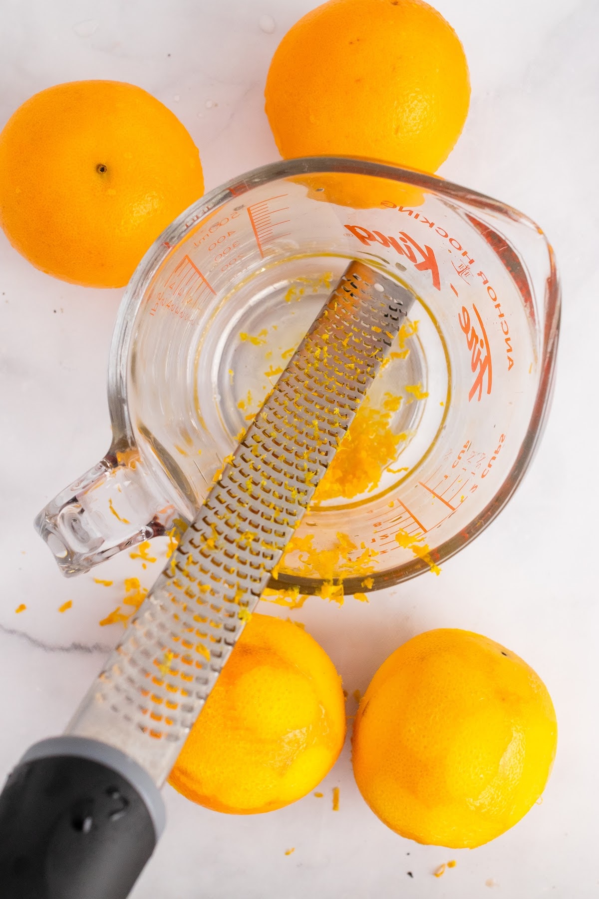 Glass measuring cup filled with orange zest with a microplane sitting on top of it with zested oranges and whole oranges around it.