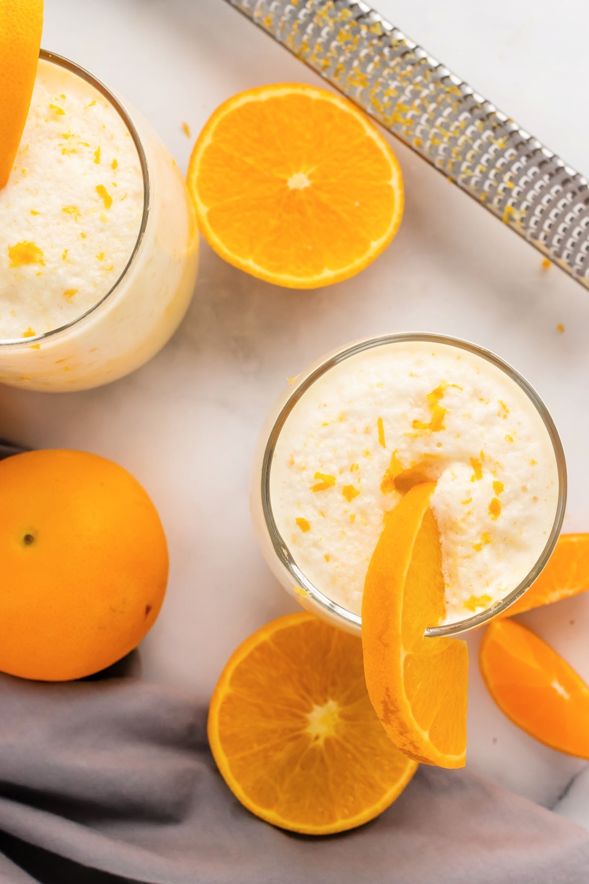 Two glasses full or a creamy orange smoothie with a microplane sitting next to them as well as oranges and orange slices.
