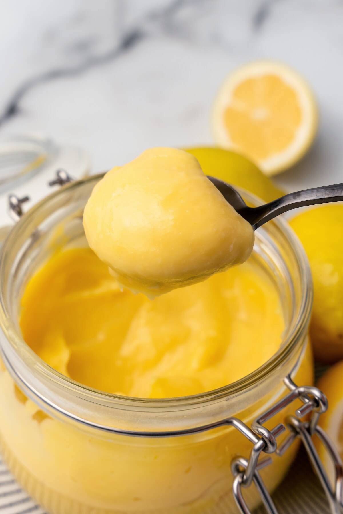 A small glass canning jar with the lid opened, filled with lemon curd sitting on a black and white striped plate with whole lemons next to it, a fresh lemon half in the background and a serving spoon full of lemon curd above the jar.