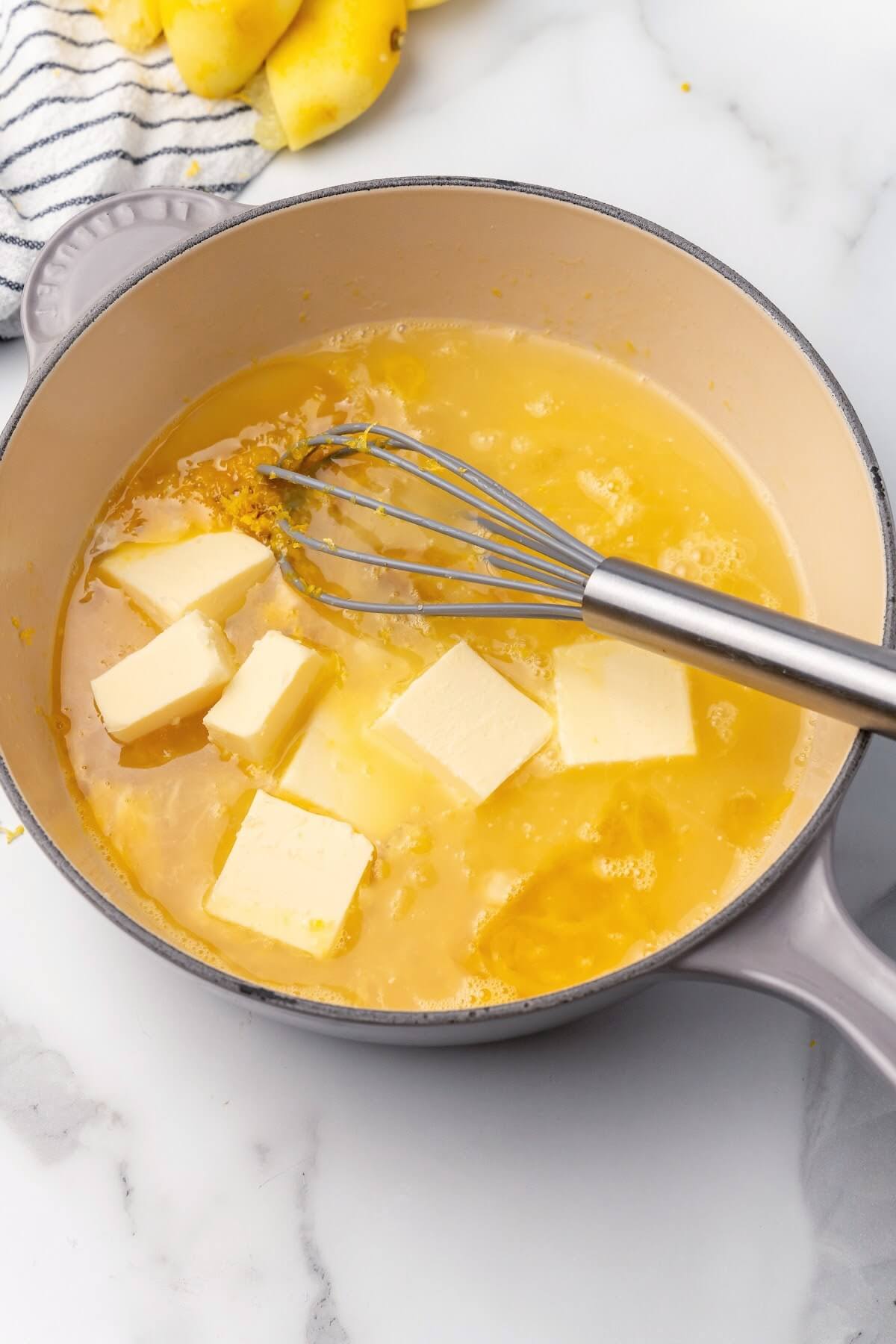 A saucepan filled with a mixture of several tablespoon pats of butter, lemon zest, lemon juice and beaten eggs with a whisk sitting on top of a marble countertop with freshly squeezed lemon halves in the background on top of a gray and white kitchen towel.