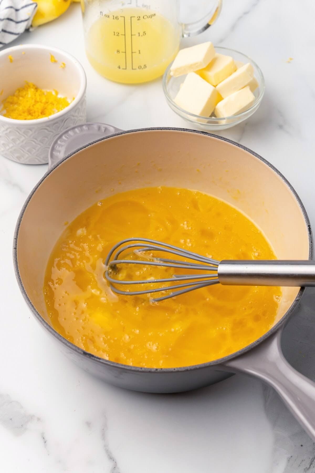 A process photo with a saucepan filled with a mixture of beaten eggs with a whisk in it sitting on top of a marble countertop with a small glass bowl filled with several tablespoon pats of butter, a glass measuring cup filled with freshly squeezed lemon juice and a small ramekin bowl with lemon zest behind the pan on the counter.