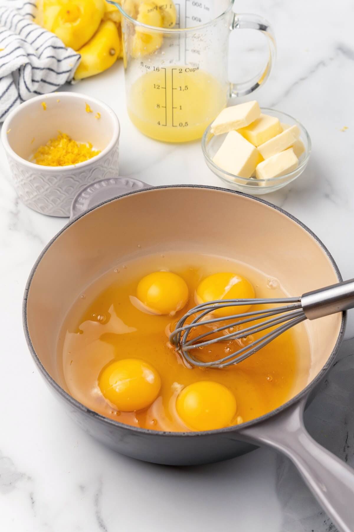 A process photo with a saucepan filled with a mixture of four eggs with a whisk in it sitting on top of a marble countertop with a small glass bowl filled with several tablespoon pats of butter, a glass measuring cup filled with freshly squeezed lemon juice and a small ramekin bowl with lemon zest, several freshly squeezed lemon halves and a gray a white striped kitchen towel behind the pan on the counter.