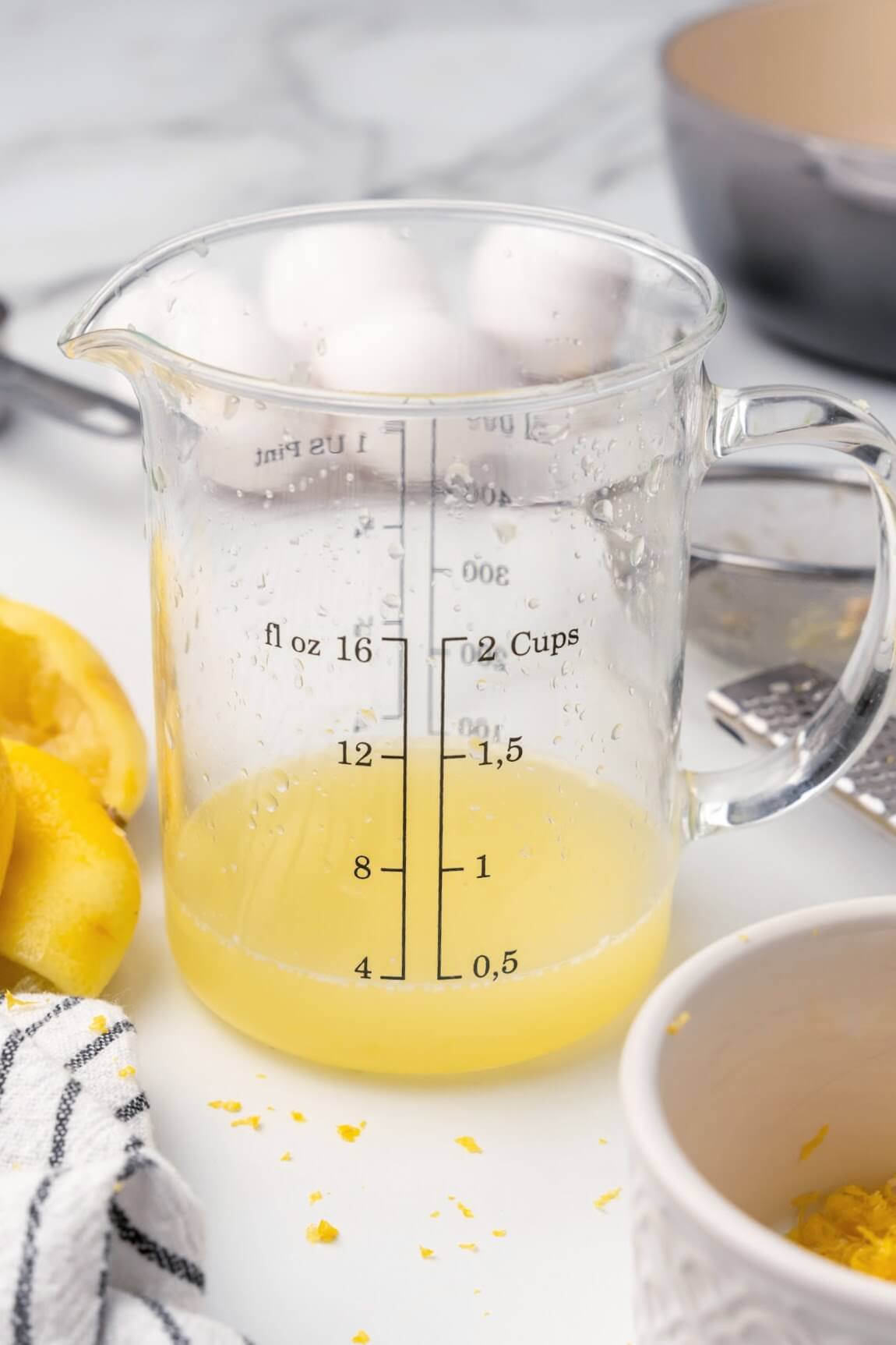 A glass measuring cup filled with ½ cup of freshly squeezed lemon juice with freshly squeezed lemon halves and a gray and white striped kitchen towel sitting beside it, a small ramekin with some fresh lemon zest in front of it and 4 whole eggs, a fine mesh strainer, a lemon microplane zester and a saucepan in the background.