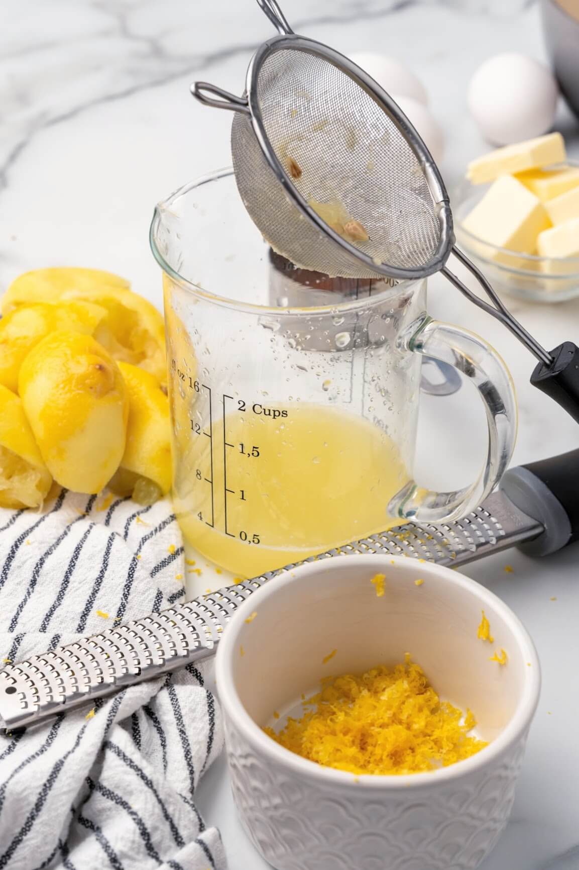 A glass measuring cup with a fine mesh strainer on top of it filled with ½ cup of freshly squeezed lemon juice with freshly squeezed lemon halves and a gray and white striped kitchen towel sitting beside it, a small ramekin with some fresh lemon zest and a microplane zester in front of it and 4 whole eggs and a small glass bowl with several tablespoon pats of butter in the background.