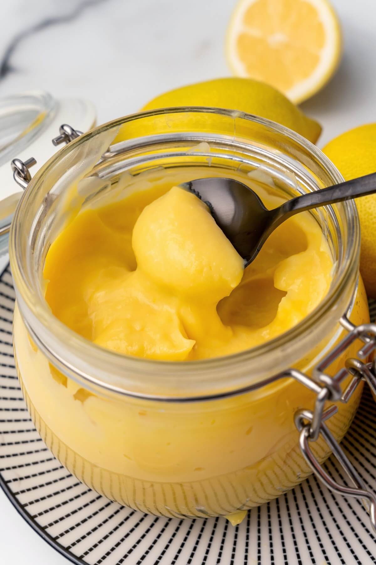 A small glass canning jar with the lid opened, filled with lemon curd sitting on a black and white striped plate with whole lemons next to it, a fresh lemon half in the background and a serving spoon full of lemon curd above the jar.