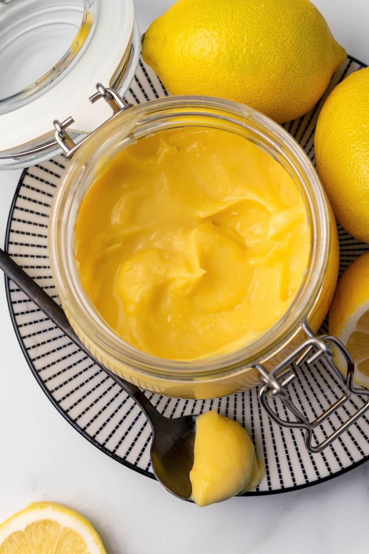 An overview view of a small glass canning jar with the lid opened, filled with lemon curd sitting on a black and white striped plate with fresh whole and halved lemons next to it and a serving spoon full of lemon curd sitting on the plate next to the jar.