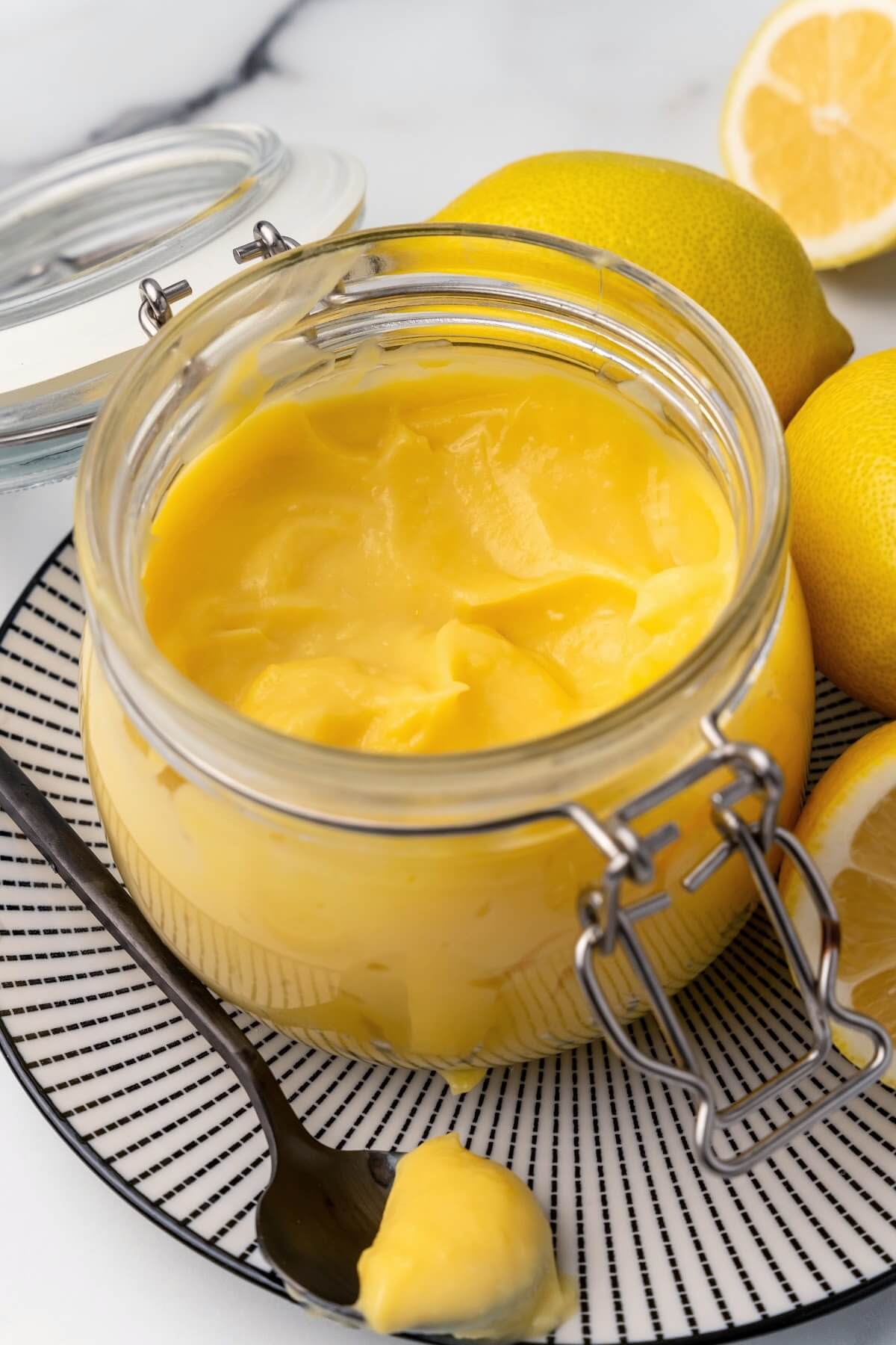 A small glass canning jar with the lid opened, filled with lemon curd sitting on a black and white striped plate with whole lemons next to it, a fresh lemon half in the background and a serving spoon full of lemon curd sitting on the plate next to the jar.