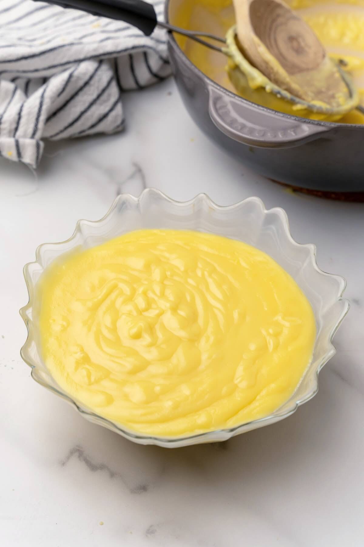 A large glass bowl full of lemon curd sitting on a marble countertop with a saucepan behind it with the remnants of the cooked lemon curd, a wooden spoon and a fine mesh strainer sitting in the pan with a gray a white striped kitchen towel sitting next to it.