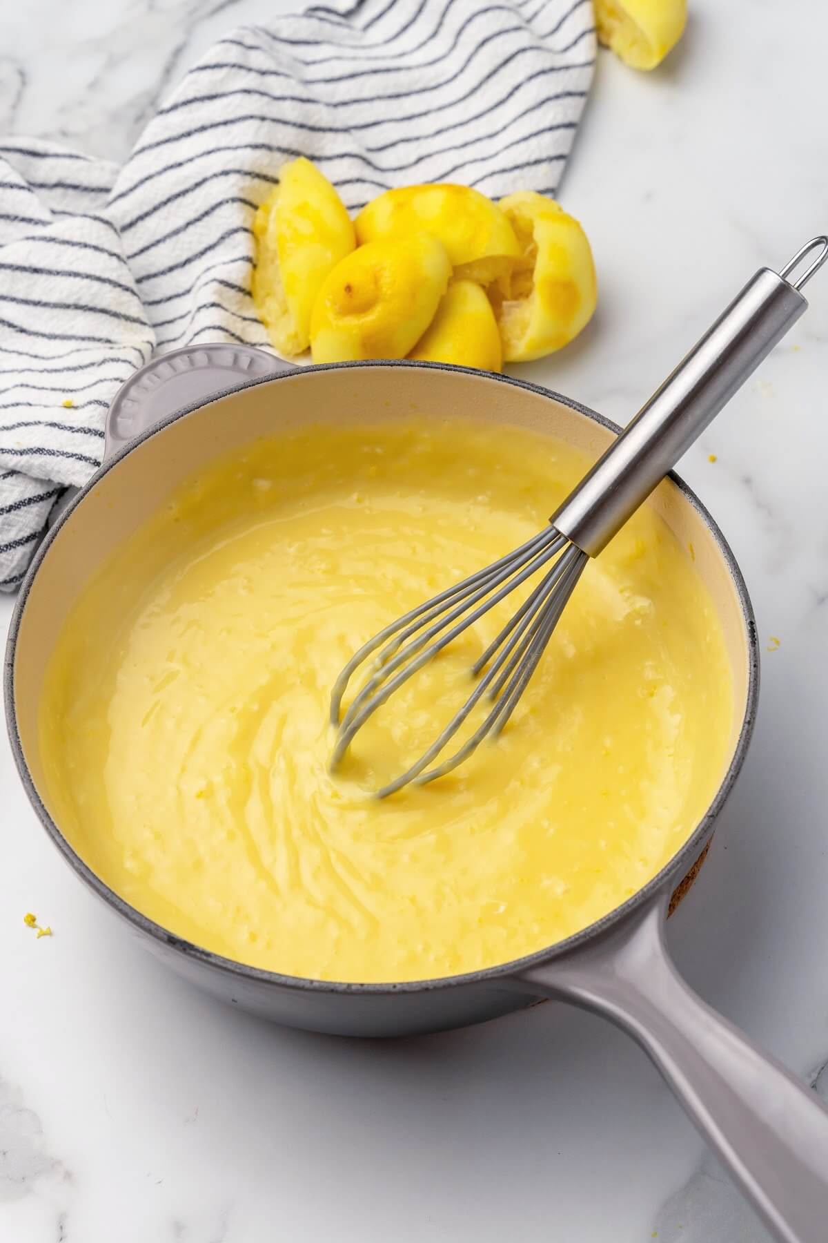 A saucepan full of lemon curd with a whisk sitting inside it on top of a marble countertop with freshly squeezed halved lemons in the background sitting on top of a gray and white striped kitchen towel.