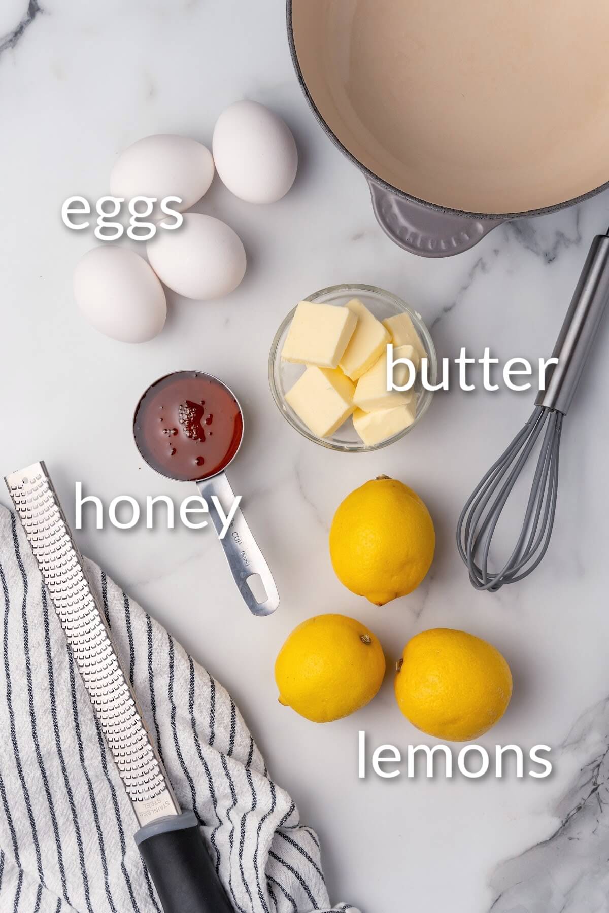 An overhead view of an empty saucepan, a whisk, four whole eggs, a small glass bowl filled with several tablespoon pats of butter, a measuring cup full of honey, 3 whole fresh lemons, a microplane zester and a gray and white kitchen towel sitting on top of a marble countertop with text that says, eggs, butter, honey, lemons.