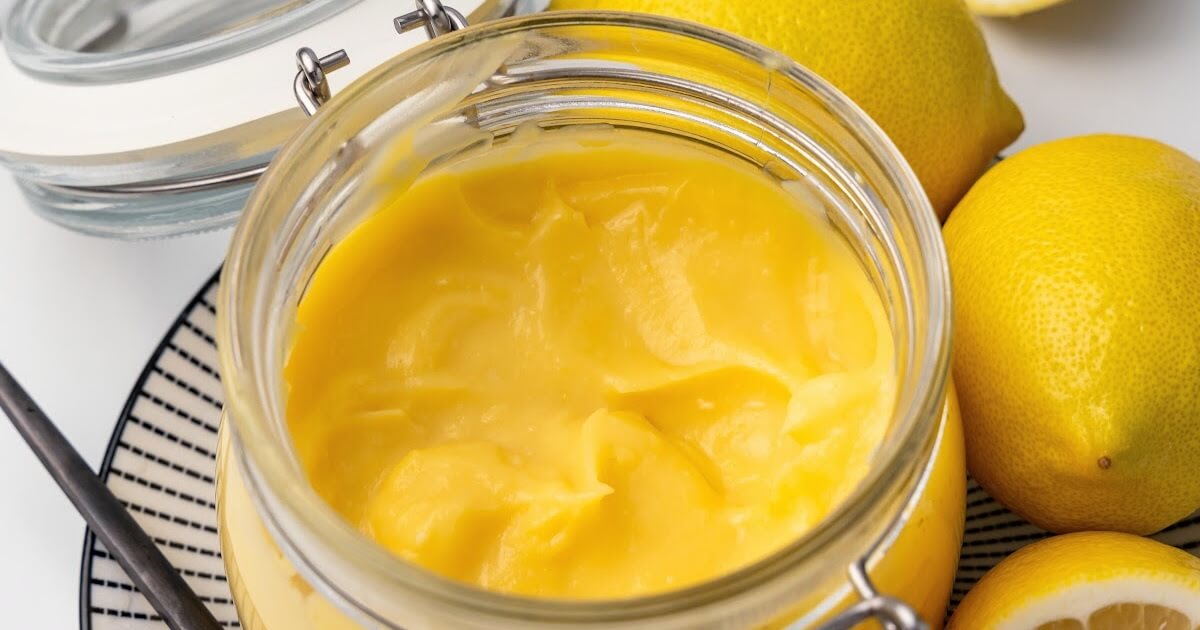 Small glass canning jar with the lid opened filled with lemon curd sitting on a black and white striped plate with whole lemons and a serving spoon next to it.