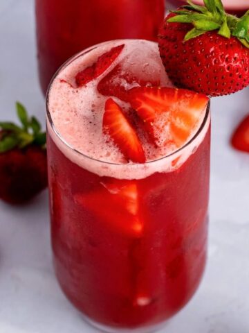 A closeup view of a glass filled with a red icy drink garnished with sliced strawberries inside of it and a fresh strawberry on the rim of the glass, sitting on top of a marble surface with fresh whole and sliced strawberries sitting around it and another glass filled with the red drink in the background all sitting on a marble surface.