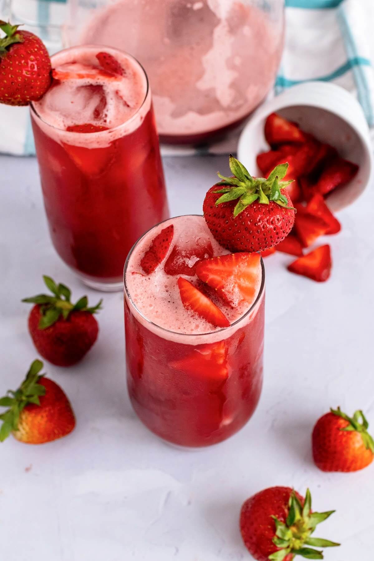 Two tall glasses filled with an icy red drink, garnished with fresh strawberry slices inside the glasses and a fresh strawberry on the rim of the glasses with fresh strawberries sitting around them, a white glass ramekin tipped over filled with strawberry slices and a glass pitcher ¼ filled with the red drink in the background sitting on top of a blue and white striped kitchen towel on top of a marble surface.