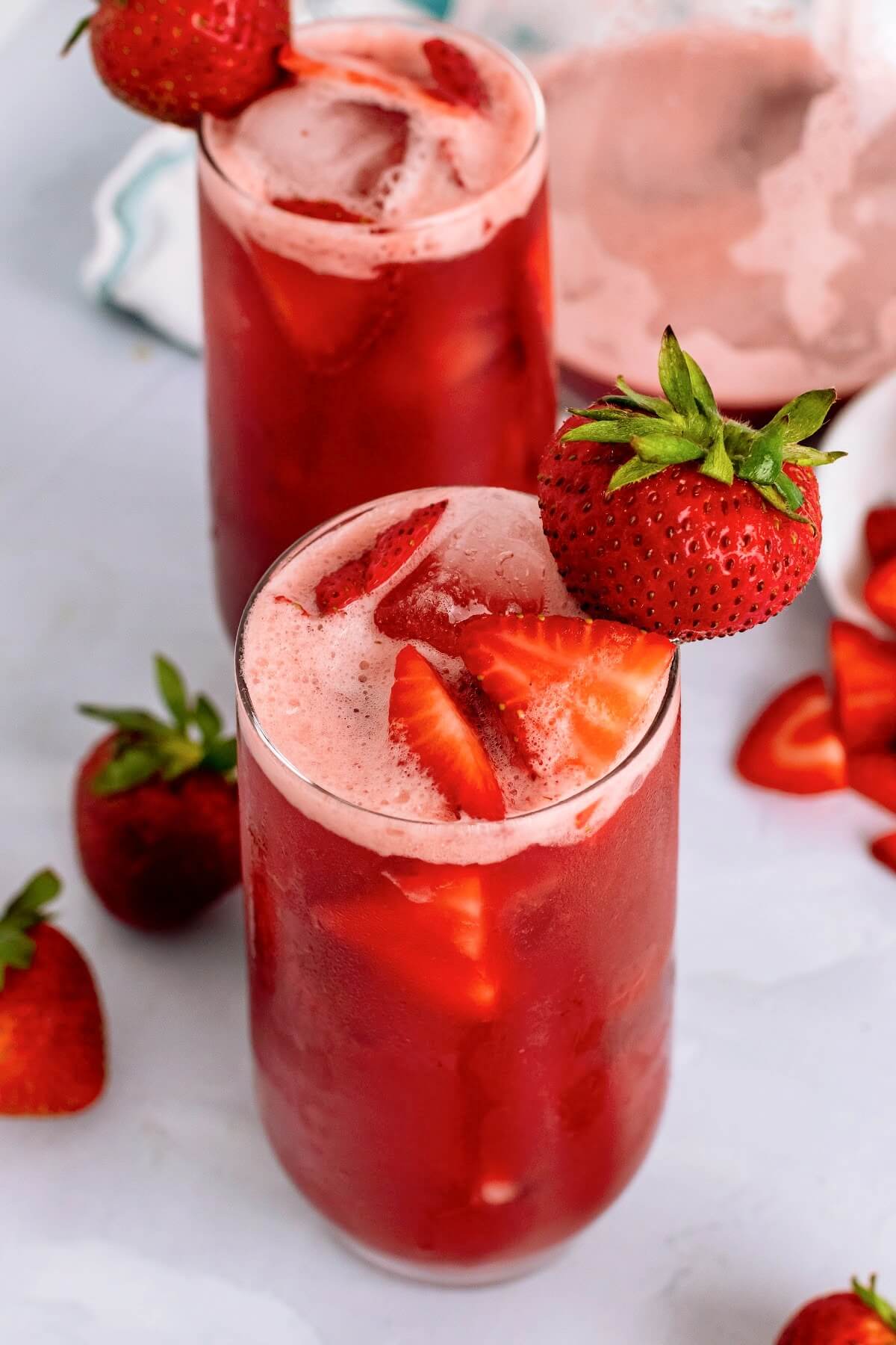 Two tall glasses filled with an icy red drink, garnished with fresh strawberry slices inside the glasses and a fresh strawberry on the rim of the glasses with fresh strawberries sitting around them, a white glass ramekin tipped over filled with strawberry slices and a glass pitcher ¼ filled with the red drink in the background sitting on top of a blue and white striped kitchen towel on top of a marble surface.