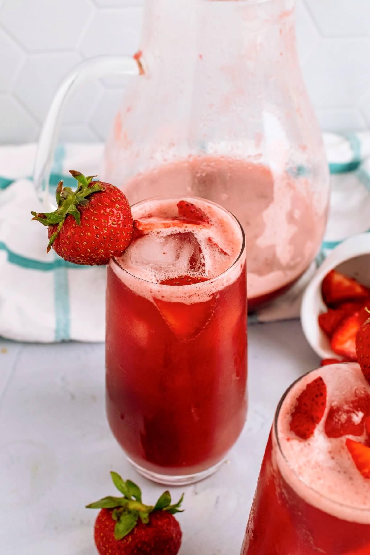 A tall glass filled with an icy red drink, garnished with fresh strawberry slices inside the glass and a fresh strawberry on the rim of the glass with fresh strawberries sitting around it with another filled glass sitting in front of it and a white glass ramekin tipped over in the background filled with strawberry slices and a glass pitcher ¼ filled with the red drink in the background sitting on top of a blue and white striped kitchen towel on top of a marble surface.