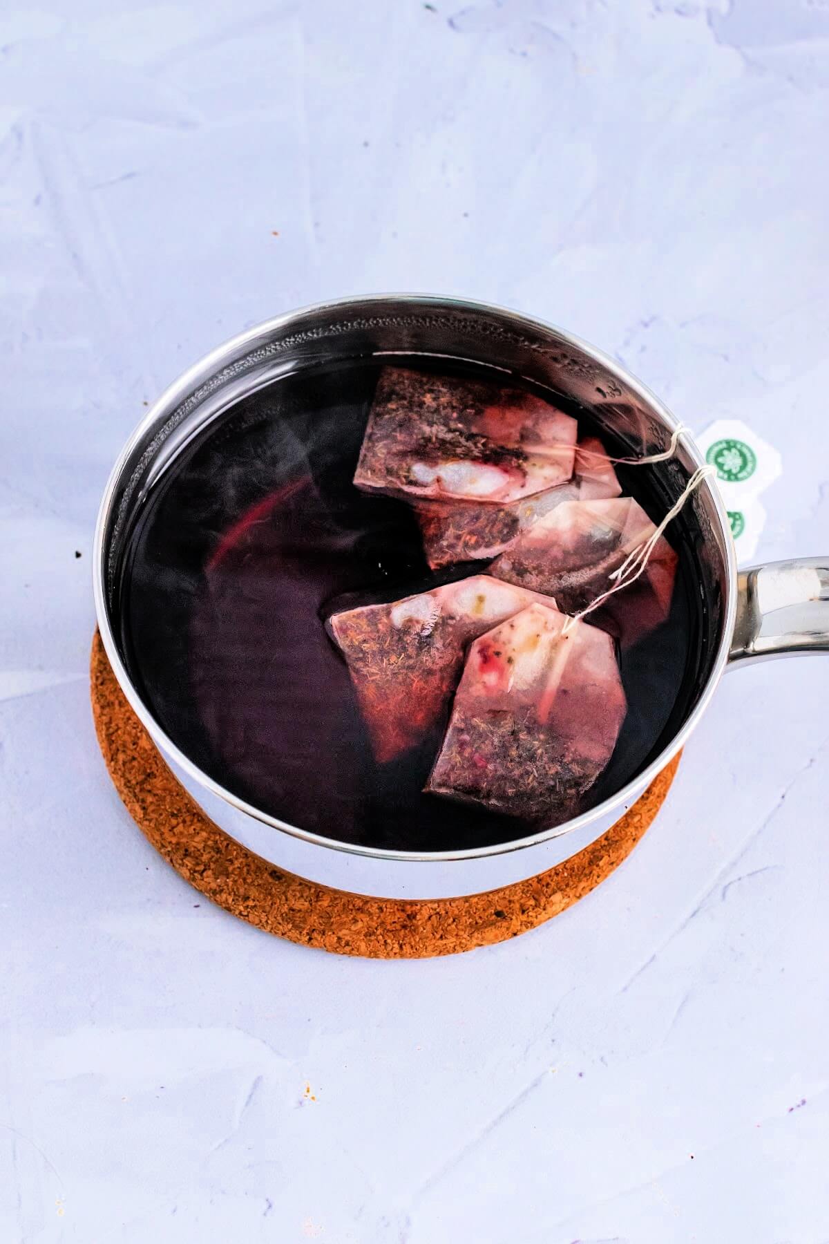 An overhead photo of a stainless steel pan filled with water and several tea bags that have steeped into the water making a red colored tea, sitting on top of a cork trivet on top of a marble surface.
