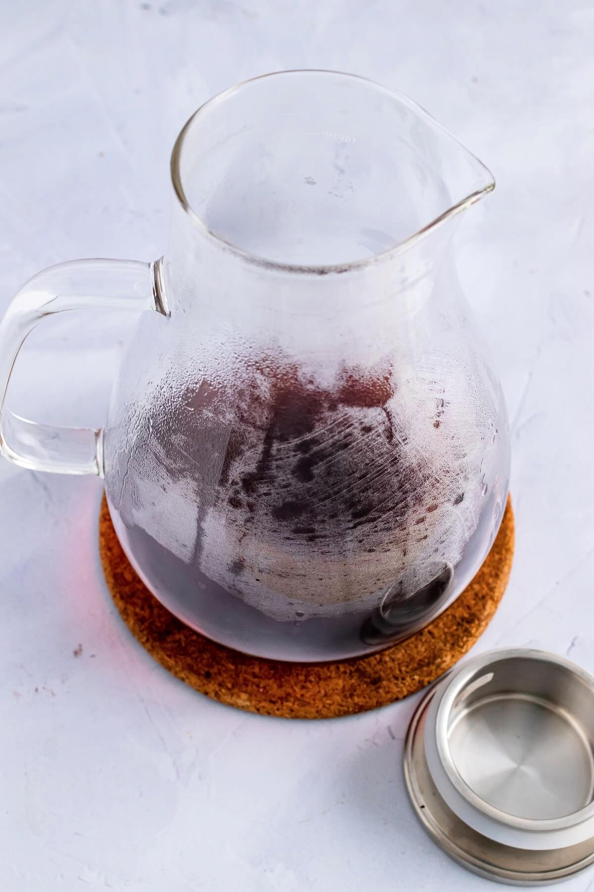 A photo of a glass pitcher ¼ filled with a hot red colored tea, sitting on top of a cork trivet on top of a marble surface with a silver lid for the pitcher sitting next the pitcher.