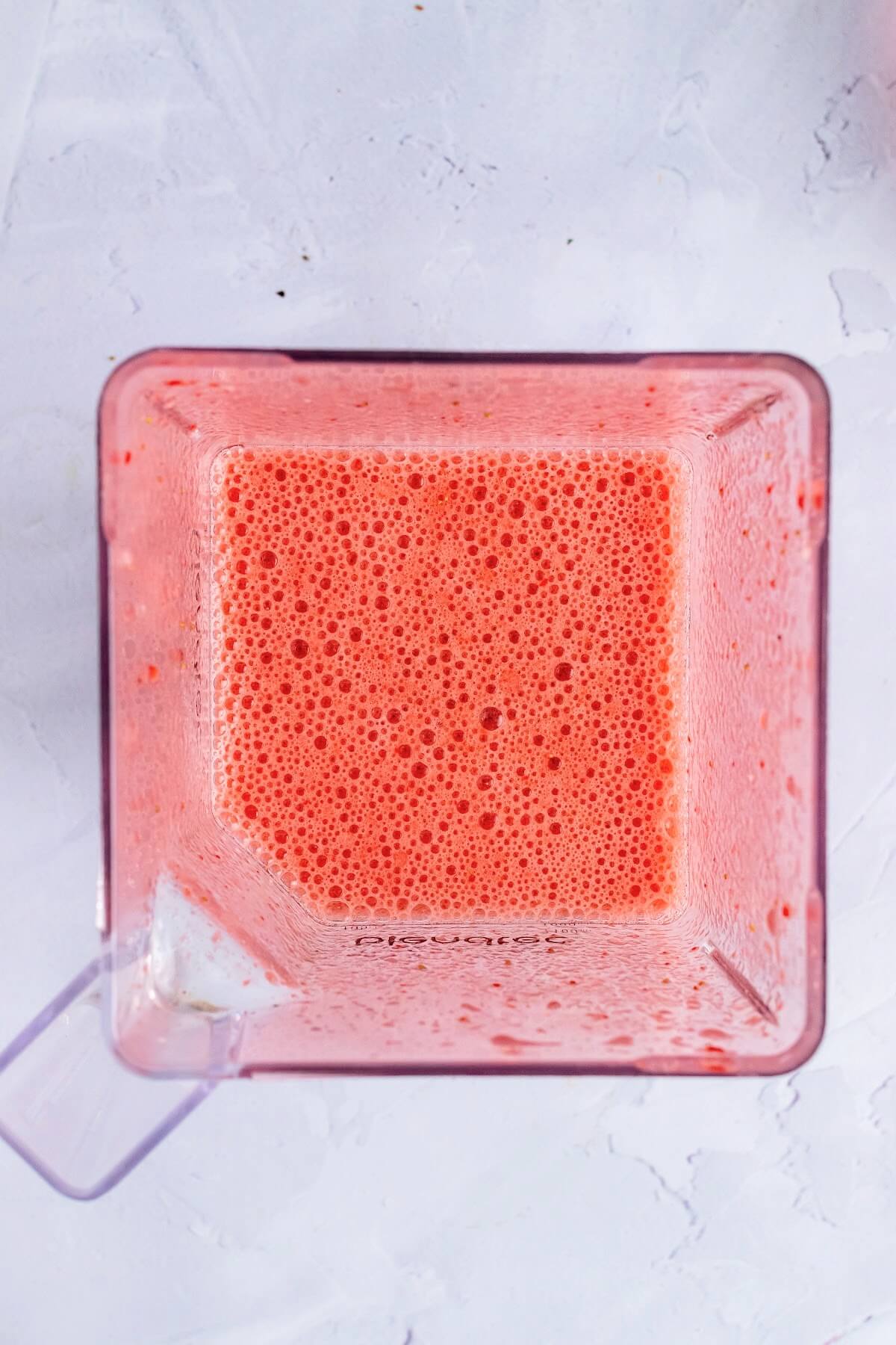 An overhead view of a blender filled with a blended, frothy red liquid, sitting on top of a marble surface.