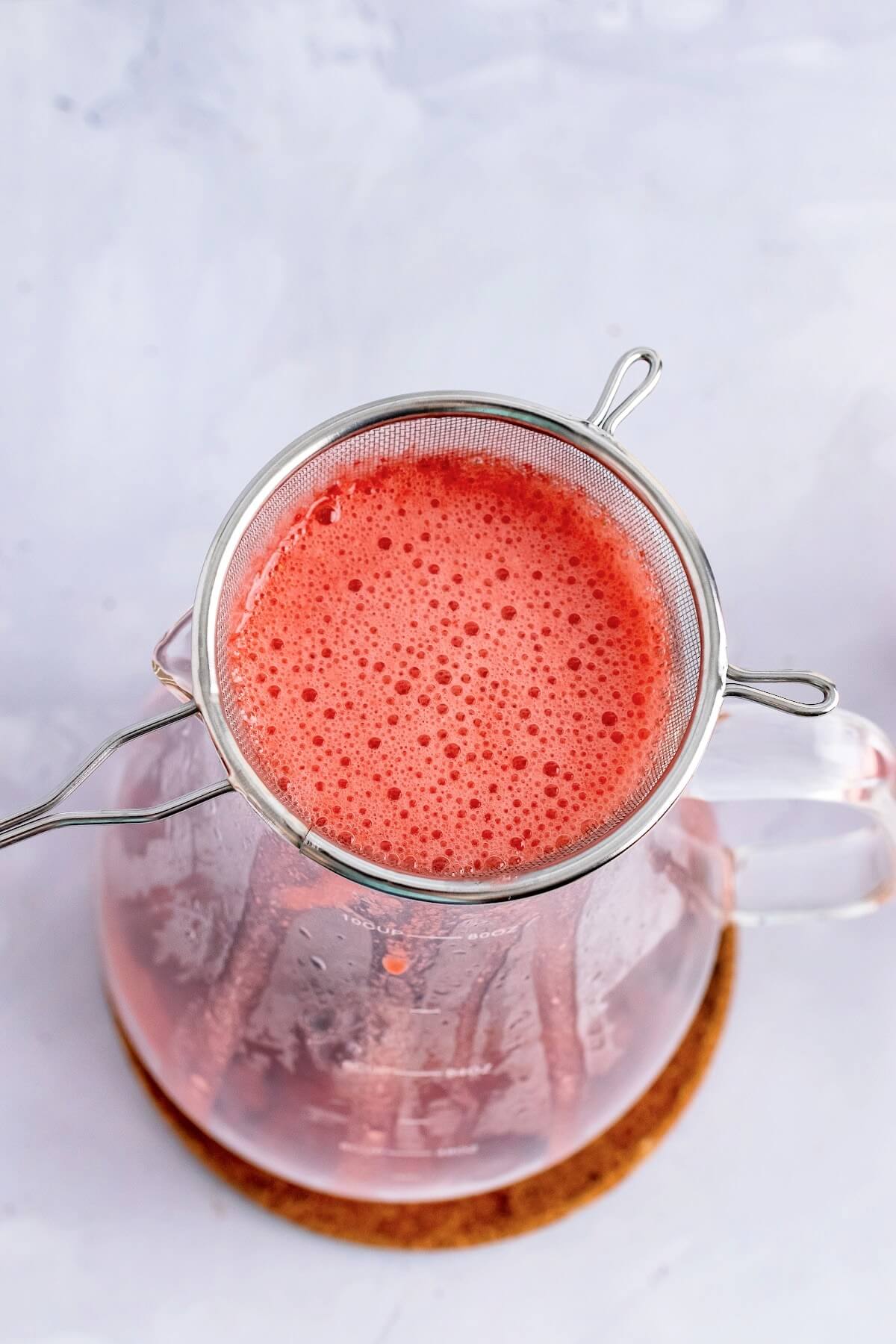 An overhead view of a fine mesh strainer sitting on top of a glass pitcher with a frothy red liquid in the sieve strainer draining into the pitcher, sitting on top of a cork trivet that's on top of a marble surface.