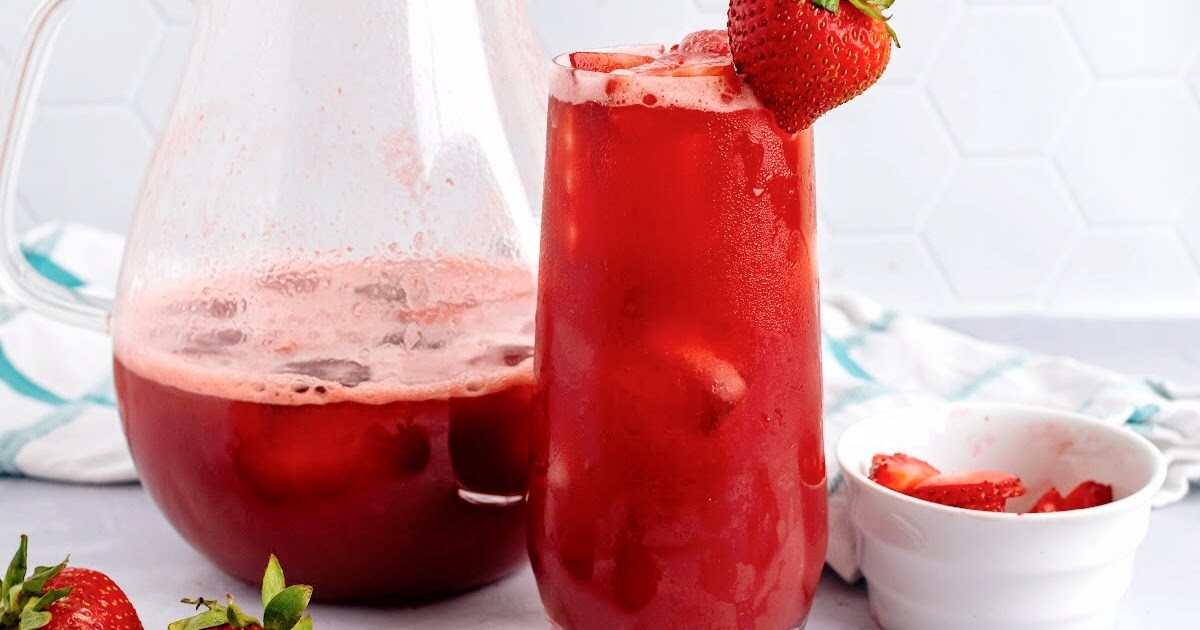 A tall glass filled with an icy red drink, garnished with fresh strawberry slices inside the glass and a fresh strawberry on the rim of the glass with fresh strawberries sitting around it, a small white glass bowl filled with strawberry slices to the right side of the glass and a glass pitcher ¼ filled with the red drink sitting to the left side with a blue and white striped kitchen towel behind it sitting on a marble surface and a white tile backdrop.