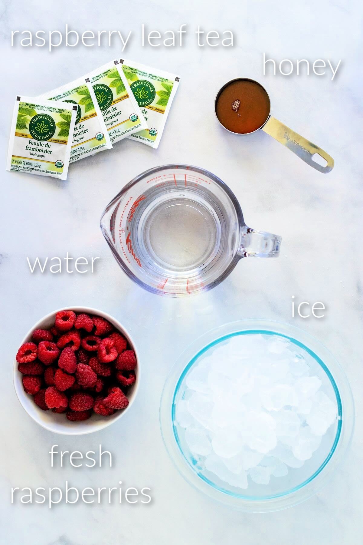 Ingredients for homemade raspberry sweet tea: a glass bowl of ice, glass measuring cup of water, measuring cup of honey, a white bowl filled with fresh raspberries, and four red raspberry leaf tea bag packages. Text overlay reads: ice, fresh raspberries, water, honey, and red raspberry leaf tea.