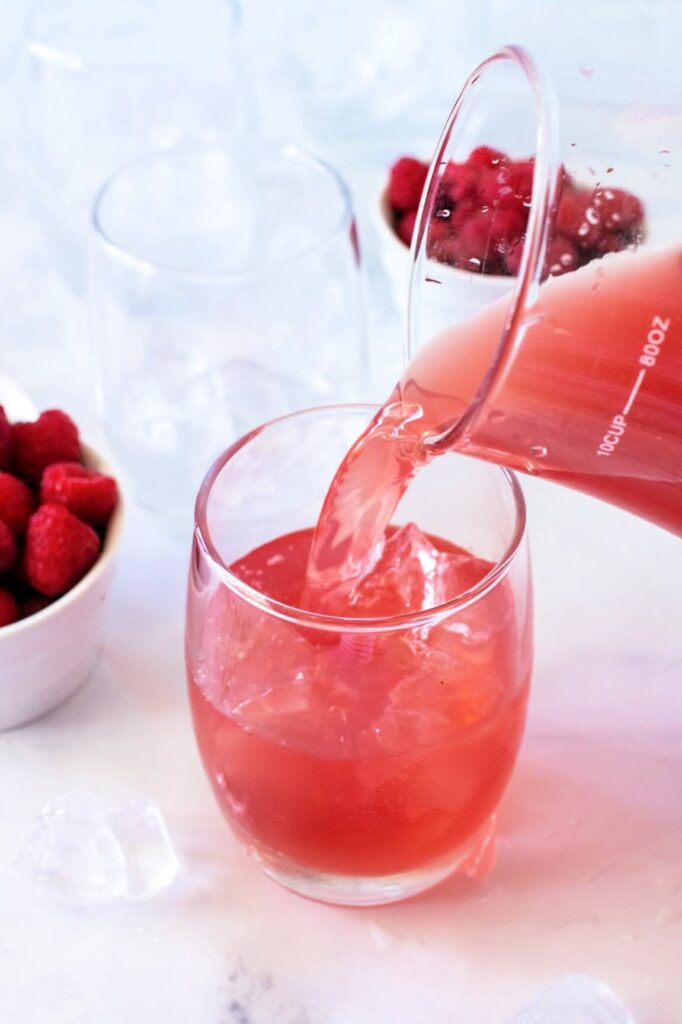 Glass pitcher pouring raspberry tea into a glass filled with ice, surrounded by two additional glasses filled with ice and two white bowls of fresh raspberries, all set on a white and gray marble surface with scattered ice cubes.