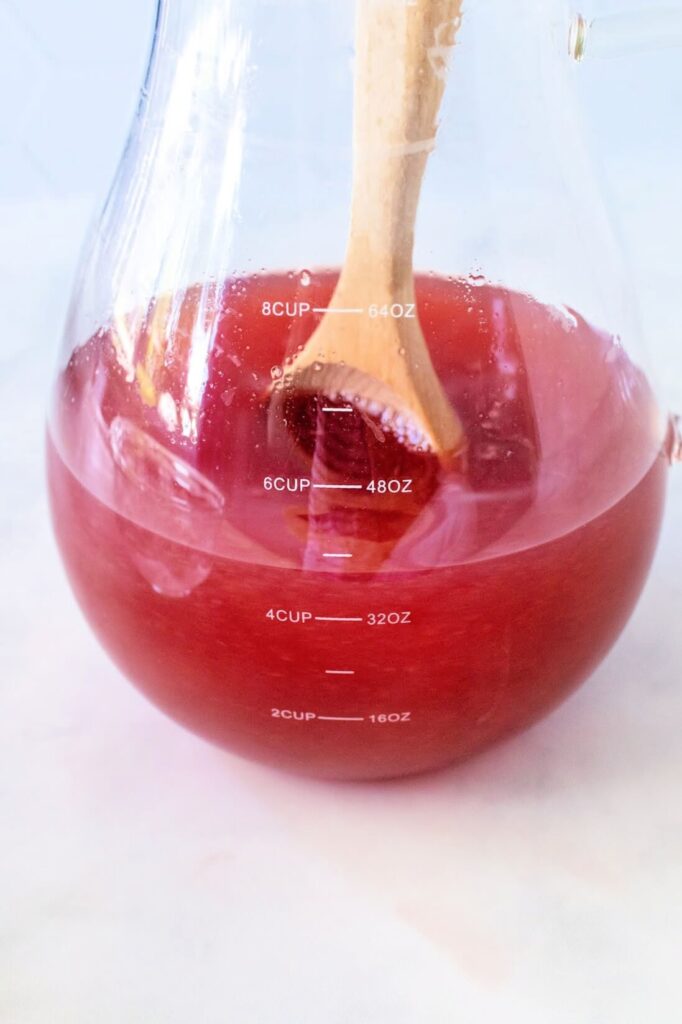 Glass pitcher filled with over 4 cups of raspberry tea mixture, stirred with a wooden spoon, sitting on a white and gray marble countertop.
